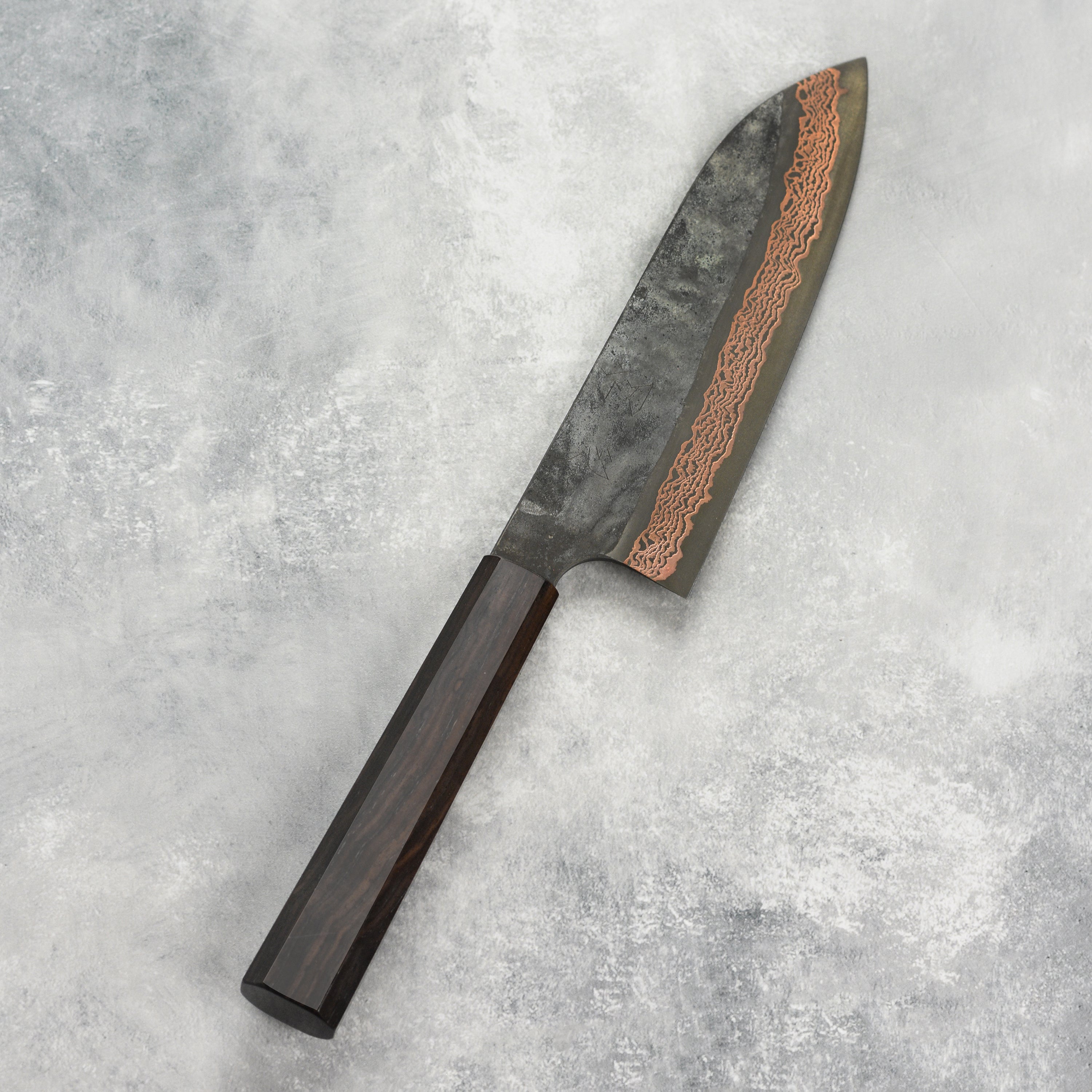 Close-up of the Hatsukokoro Santoku Knife on a wooden cutting board, highlighting its copper Damascus finish.
