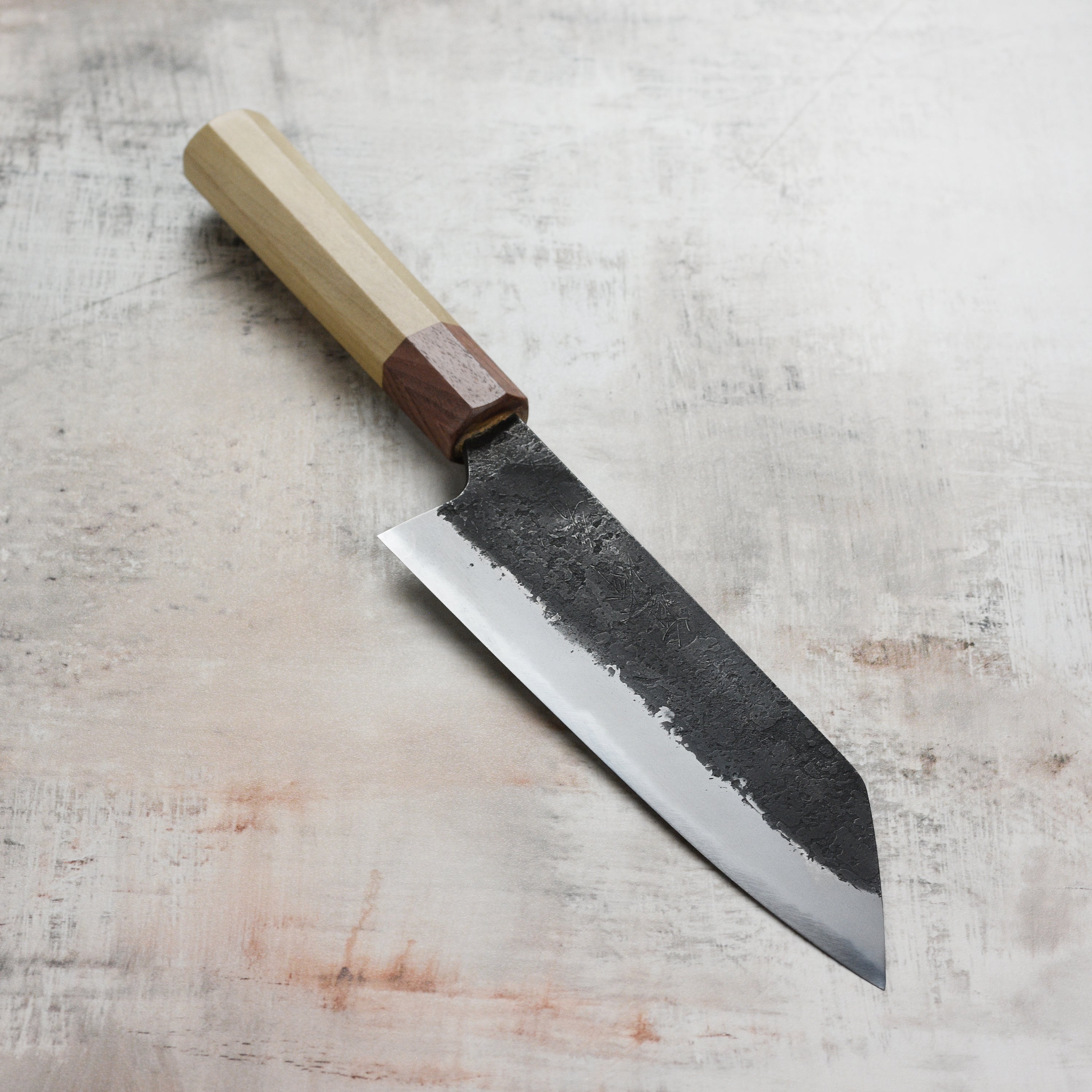 Mazaki Shirogami Santoku knife displayed with assorted herbs, showcasing its precision cutting edge.