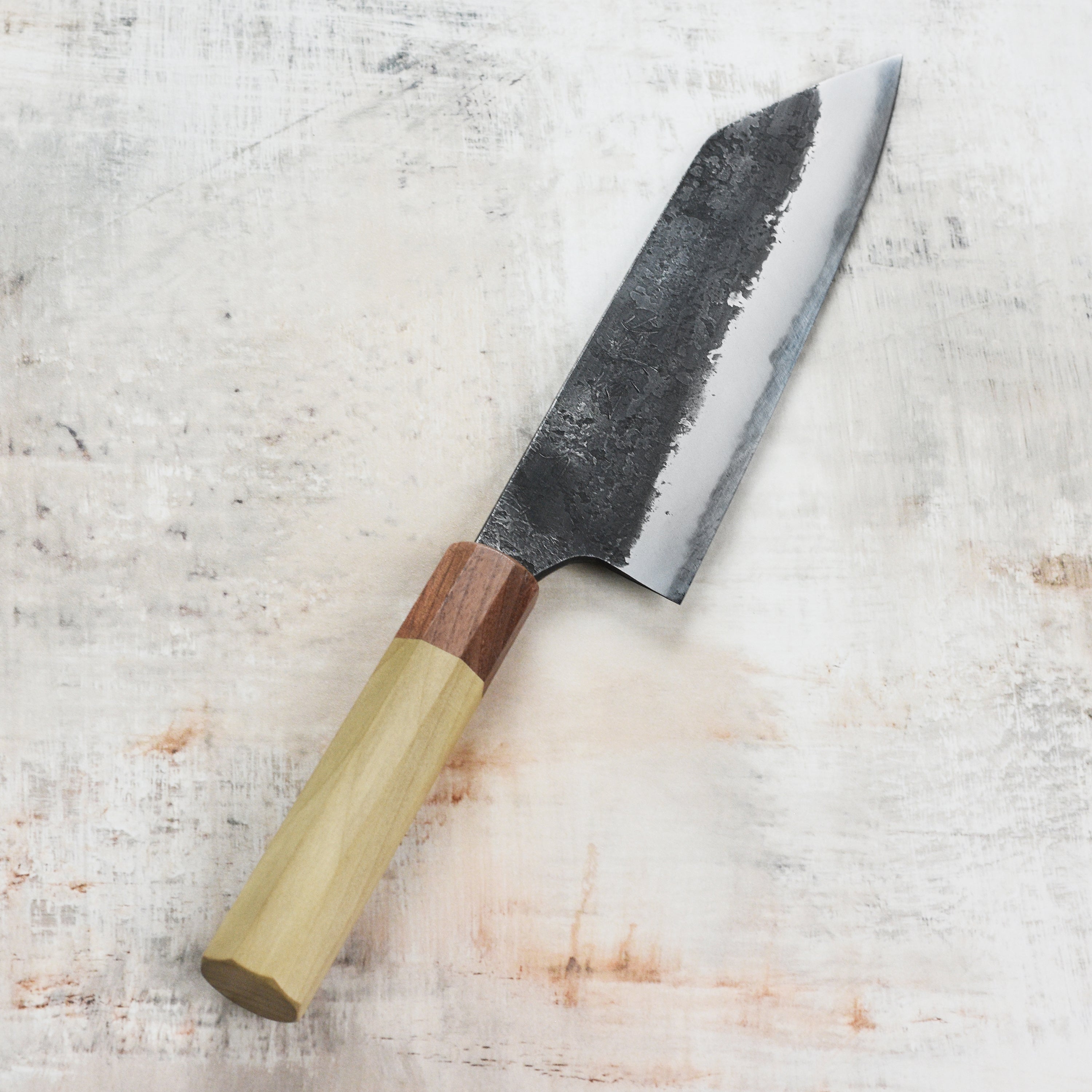 Mazaki Shirogami Santoku knife displayed with assorted herbs, showcasing its precision cutting edge.