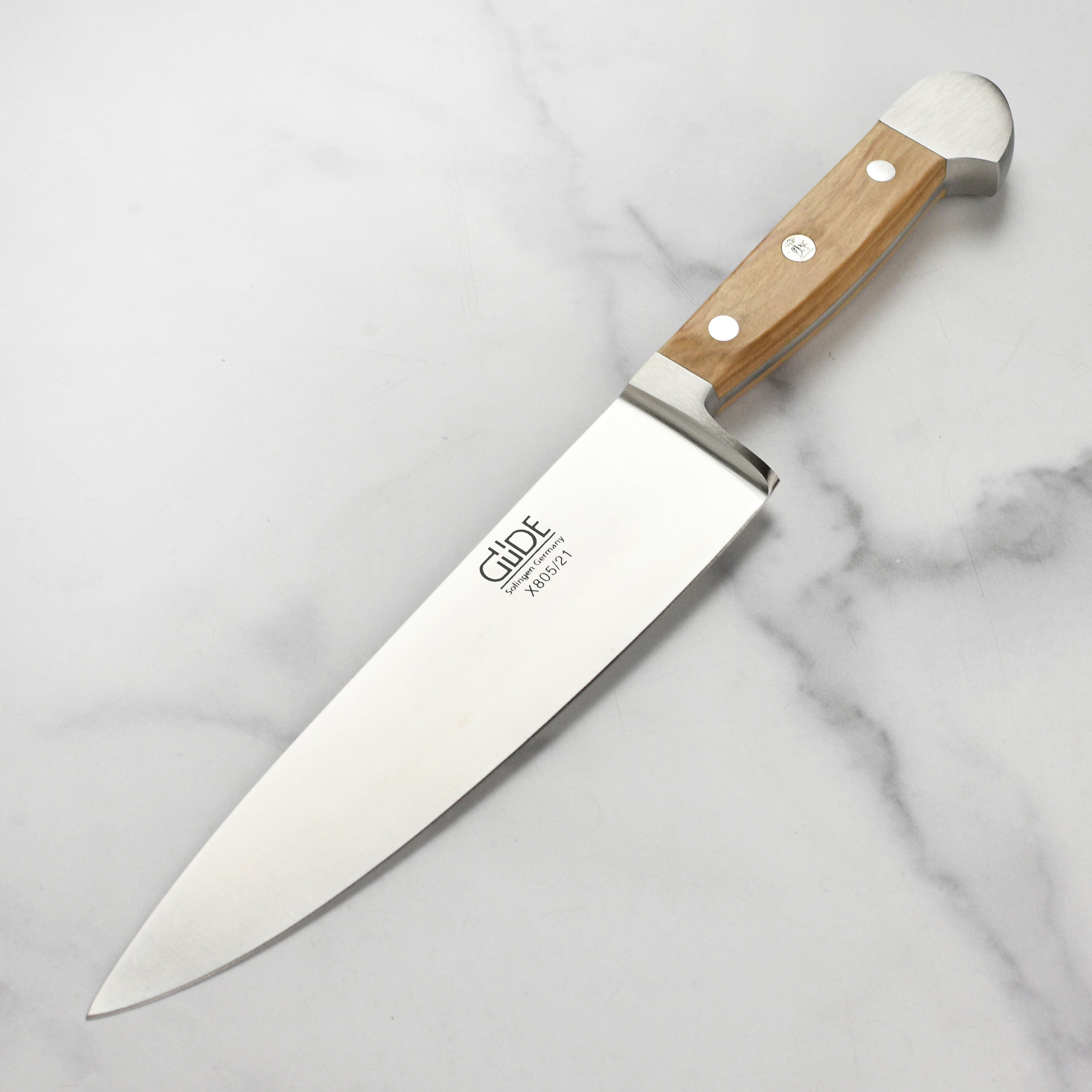 Gude Alpha Olive Chef's Knife displayed on a wooden cutting board, highlighting its ergonomic olive wood handle.