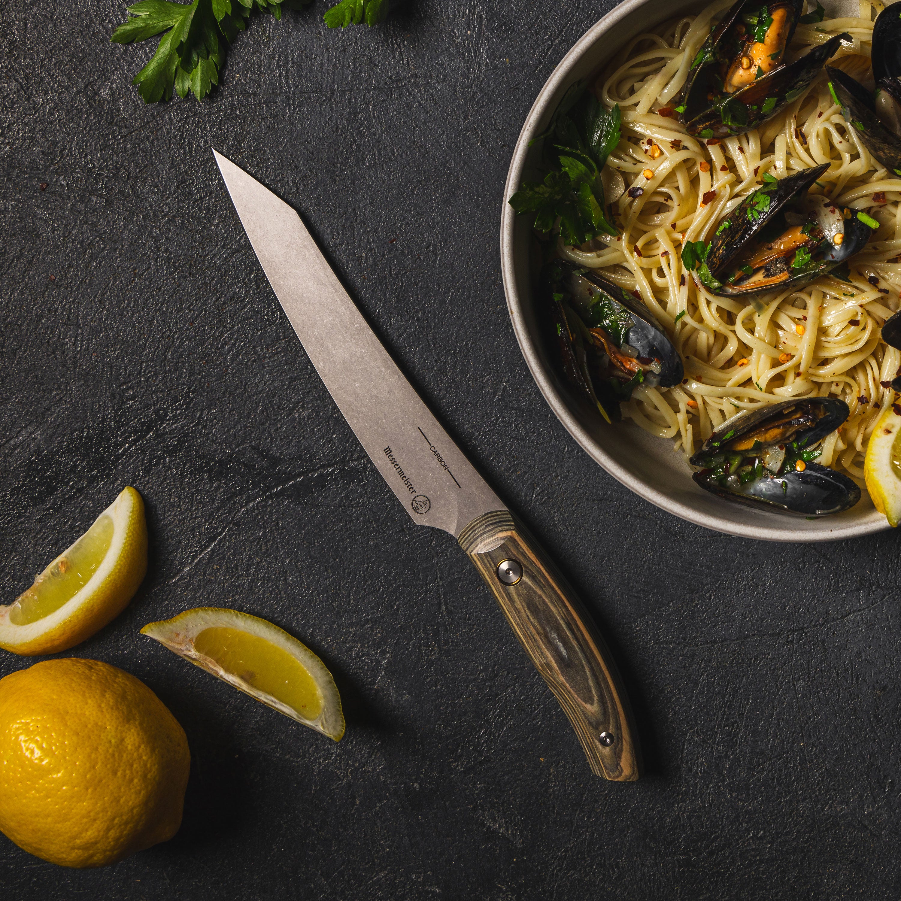 Messermeister Artisan Carbon Utility Knife displayed on a wooden cutting board emphasizing its handcrafted design.